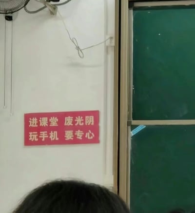 这是一张教室内的实拍画面，墙面左上角悬挂着一个风扇，右侧是教室的绿色黑板，墙面上贴着一张红底白字的标语，内容为“进课堂 废光阴 玩手机 要专心”，该标语是对传统劝学标语的反向调侃，将正常的逻辑颠倒，用反讽的方式吐槽部分学生在课堂上无心学习、沉迷手机的状态，画面下方露出一个人的头顶，整体场景还原了真实的教室环境，充满趣味和调侃意味。