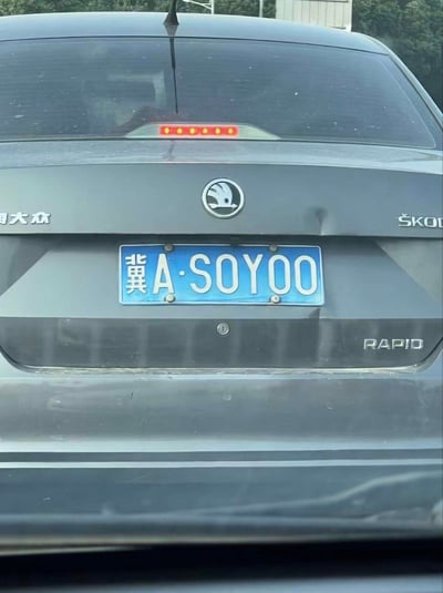 这是一张道路上随手拍摄的上汽大众斯柯达RAPID车型的车尾照片，车辆悬挂河北石家庄（冀A）的蓝底白字机动车号牌，车牌号为冀A·SOYOO，字母数字组合谐音英文短语“SO YOU”，自带趣味效果。车尾可见斯柯达品牌标识、“RAPID”车型标识、“上汽大众”字样，高位刹车灯处于熄灭状态，车身附着一些灰尘，显得有些脏污。