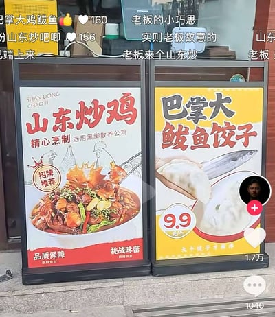 这是一张社交媒体截图，内容是餐馆门口并排摆放的两块菜品宣传立牌，左侧立牌宣传山东炒鸡，标注了选用黑脚散养公鸡，为招牌推荐，承诺品质保障，配有炒鸡成品菜品图；右侧立牌宣传9.9元每个的巴掌大鲅鱼饺子，配有鲅鱼和饺子的示意图。由于两块立牌相邻，拼接后的文字连读会产生不雅谐音梗，图中还带有社交媒体的点赞、评论互动数据，以及网友的评论，网友调侃这是老板故意设计的小巧思。