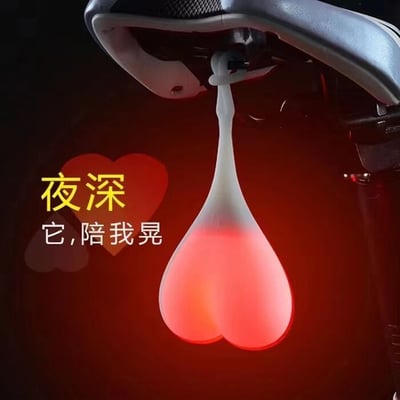 图片展示了一个安装在自行车座椅下的红色心形LED灯，灯光在深色背景下显得格外醒目。灯的形状设计成爱心样式，随着骑行会自然晃动。图片左侧配有黄色和白色文字“夜深 它,陪我晃”，结合心形灯的设计，形成了一种拟人化的幽默效果，暗示这个灯在夜晚骑行时成为了陪伴者。