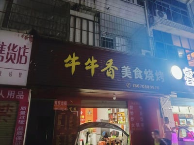夜晚的街道场景，中心是一家名为“牛牛香美食烧烤”的店铺，招牌为深色底黄色大字，下方标注联系电话18670689976。店铺门口亮着灯，可见内部陈设，门口贴有红色“有房出租”招贴（附电话18692338135）。左侧相邻店铺有“杂货店”“生活馆”等招牌，右侧有其他商铺灯光。背景为带有铁窗和空调外机的老旧居民楼，整体呈现出浓厚的市井生活气息，街道上有行人活动，烟火气十足。