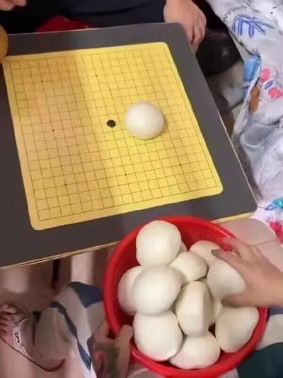 一张俯拍视角的照片，展示了一个有趣的生活场景。画面中央是一块黄色网格的围棋棋盘，棋盘上除了一颗黑色棋子外，还放着一个完整的白色馒头，仿佛被当作巨型棋子使用。棋盘下方有一个红色的塑料篮子，里面装满了多个大小相似的白馒头，一只手正伸进篮子里准备拿取。背景中可以看到几个人的手脚和花色的衣物，营造出一种家庭聚会或朋友间轻松玩乐的氛围。