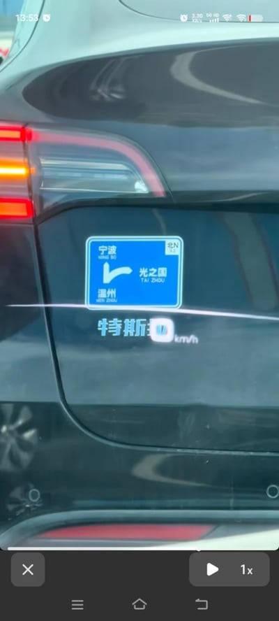 这是一张视频截图，内容为黑色汽车的车尾局部，车尾处贴有模仿国内高速交通指示牌样式的蓝色趣味车贴，车贴按照正规路牌的格式标注了宁波、温州两个地名，箭头指向的目的地被恶搞为奥特曼设定中的“光之国”，同时标注了拼音TAI ZHOU，是玩“台州”的谐音梗，画面底部可以看到手机端视频播放的控制按钮，说明该截图截取自短视频内容。