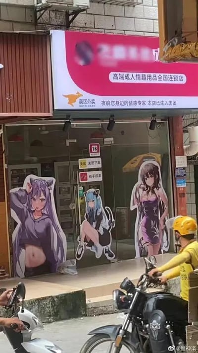 这是一张街头实拍的竖版照片，画面主体是一家名为“高端成人情趣用品全国连锁店”的店铺，店铺招牌为粉色，标注了已加入美团外卖，标语为“夜疯您身边的情感专家”。店铺的玻璃门上贴有三张风格软萌性感的二次元美少女立牌贴纸，分别是穿露脐装的紫色长发少女、蹲坐姿态的蓝发少女、身着紧身裙的棕色长发少女，这种二次元萌系风格和成人用品店的定位形成强烈的违和感。画面下方有两位骑电动车的路人，其中一位是美团外卖员，进一步强化了店铺和美团的关联。