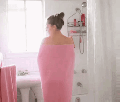 这是一个趣味动态GIF，画面中一位扎丸子头、戴眼镜的女生身处浴室，身上裹着粉色毛巾。在她转身的过程中，手臂与毛巾的位置形成了视觉错位，看起来仿佛毛巾滑落露出了上半身，实际是视觉错觉效果，制造出荒诞搞笑的反差感，让人觉得趣味十足。