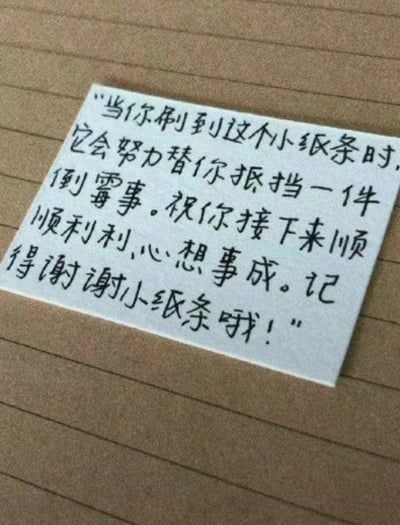图片中展示了一张白色小纸条，上面用黑色手写体写着一段幸运祝福文字，纸条被放置在棕色的横格笔记本页面上。文字内容为：“当你刷到这个小纸条时，它会努力替你抵挡一件倒霉事。祝你接下来顺顺利利，心想事成。记得谢谢小纸条哦！”整体画面温馨，传递出积极温暖的祝福氛围，旨在给看到的人带来心理慰藉和好运期盼。