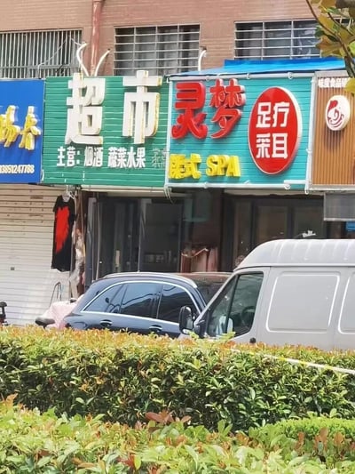 一张街头商铺照片，展示了一排临街店铺的招牌。左侧是一家蓝色招牌的店铺，中间是绿色招牌的超市，主营烟酒、蔬菜水果等商品，右侧是一家青色招牌的足疗店，名为“灵梦”，提供足疗采耳和泰式SPA服务。店铺前停放着几辆汽车，前景有绿色灌木丛。
