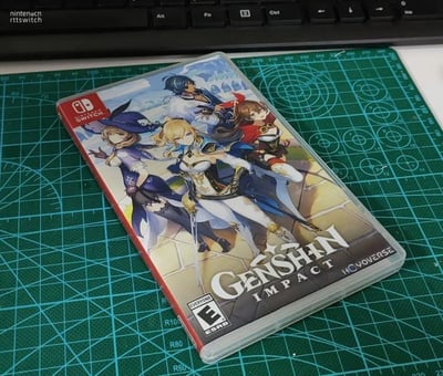 图片展示了任天堂Switch平台《原神》游戏卡带的包装盒，放置在绿色网格切割垫上，背景可见部分键盘。包装盒封面印有四位《原神》角色——旅行者、安柏、丽莎和凯亚，他们身着标志性服装，摆出自信的战斗姿态，背景为游戏中的奇幻场景。盒面左上角有Nintendo Switch标志，底部标注ESRB E级（适合所有人）评级。