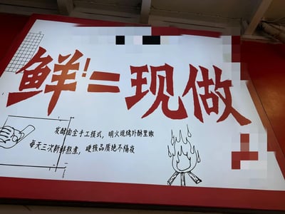 一张红色边框的白色宣传牌，中央用醒目红色大字写着“鲜=现做”。下方配有黑色小字说明：“发酵面全手工模式，明火现烤外酥里嫩，每天三次新鲜熬煮，硬核品质绝不隔夜”。左侧有手绘线条图展示手部操作食物的场景，右侧则是火焰图案，整体设计突出“新鲜现做”的食品卖点。背景疑似店铺室内环境，地面为红色，上方可见白色桌椅结构。