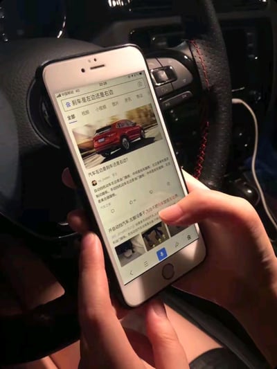 图片展示了一个人坐在汽车驾驶座上，手持智能手机搜索"刹车是左边还是右边"的场景。手机屏幕上显示着相关的搜索结果，包括一张红色SUV的图片和关于汽车刹车位置的文字说明。画面突出了新手司机可能遇到的困惑，同时也暗示了驾驶时使用手机的不安全行为。