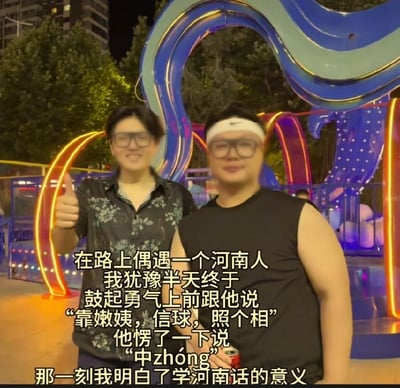 这是一张夜晚户外拍摄的真人合影梗图。画面中有两名年轻男子站在色彩鲜艳的霓虹曲线雕塑前，背景可见树木、建筑物和游乐设施。左侧男子穿黑底白花短袖衬衫、戴黑框眼镜，面带笑容并竖起大拇指；右侧男子戴白色发带和黑框眼镜，穿黑色无袖T恤，手里拿着红色饮料罐。图片下方叠加了一大段中文字幕，讲述作者偶遇河南人后，鼓起勇气用学来的河南话说“靠嫩姨，信球，照个相”，对方愣了一下后回答“中zhóng”，作者于是感慨明白了学河南话的意义。“靠嫩姨”“信球”在河南方言中属于粗俗或骂人的口头禅，正常并不适合用来礼貌请求合影；而“中”在河南话里表示“行、可以、好”。笑点在于说话者把不礼貌的方言词汇直接用在搭话中，对方却仍爽快答应，形成荒诞、反差的喜剧效果，也调侃了河南方言直爽的表达风格。