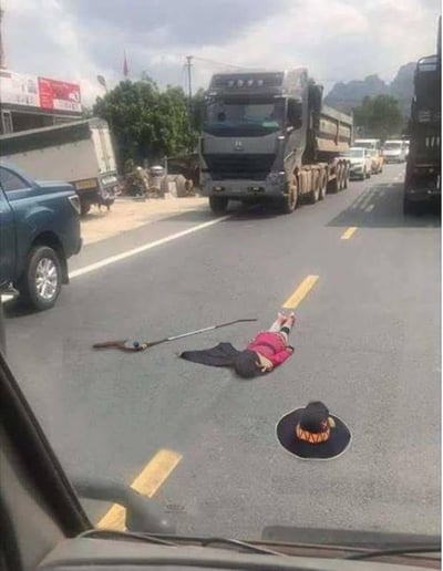 这是一张从车内视角拍摄的公路场景图，利用视觉错位制造出逼真的恶作剧效果：路面上的平面人形图案搭配放置在旁边的黑色宽檐帽、长杆道具，从车内的角度看过去，仿佛是一个穿着粉色上衣的人趴在马路中间，像是发生了意外事故。画面中还有行驶的大货车、皮卡车，路边有商铺、树木和远山，整体场景是普通的户外公路，实则是通过平面图案和道具的组合，营造出迷惑人的视觉错觉，属于趣味整活的恶作剧内容。