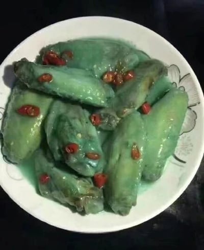 这是一张原始拍摄的静态单图，画面中展示了一盘被做成青绿色的鸡翅，鸡翅表面裹着浓稠的绿色酱汁，上面还点缀着一些切碎的红辣椒，盛放在带有浅灰色花纹的白色圆形盘子里。这道鸡翅的颜色完全不符合正常烹饪后鸡翅的色泽，属于典型的黑暗料理，大概率是烹饪过程中错误使用了食材、调料，或是和某种绿色食材发生了奇怪的反应，导致成品呈现出这种怪异的青绿色，看起来毫无食欲，充满了厨艺失误的喜感。