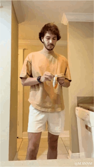 这是一段GIF动态图，画面中一位留着卷发、穿着浅棕色短袖与白色短裤的男生，身处住宅的走廊区域，正低头专注地用双手拆开手中的零食包装，场景是普通的居家环境，整体氛围日常平淡，右下角带有@M.SNNM的水印。