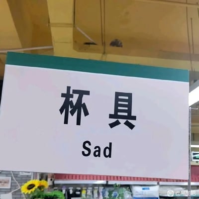 这是一张在超市内拍摄的静态实拍图，画面核心是一块带有绿色顶部的白色标识牌，标识牌上用黑色字体标注中文“杯具”，下方对应英文“Sad”。这里利用“杯具”与“悲剧”的谐音关系，将原本指代杯类餐具的词汇，用表达悲伤情绪的英文“Sad”来对应，制造出趣味十足的谐音梗效果。标识牌下方可见超市的货架、各类商品，还有黄色向日葵装饰，背景是超市的黄色天花板与货架区域，图片右下角带有“@吊图吧”的水印，整体场景充满生活化的搞笑氛围。