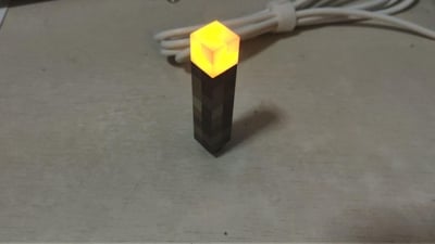 这是一张实拍照片，展示了还原沙盒游戏《我的世界》中像素火把的实体周边摆件，它被放置在浅色桌面，顶部的方块部分正发出暖黄色的光，背景可见一束捆扎的白色充电线，完美复刻游戏里火把的像素风格外观，是充满游戏情怀的桌面小装饰。