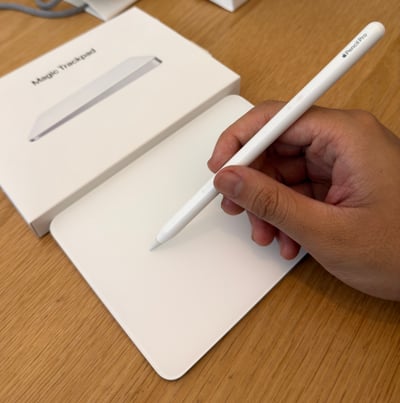 这张图展示了一只手握着Apple Pencil Pro，将笔尖放在苹果Magic Trackpad（妙控板）的表面，仿佛是在使用数位板进行创作。图中还能看到妙控板的包装盒，整体场景设置在木质桌面上，这个画面呼应了发布者“为什么苹果不出一个数位板”的想法，用现有的苹果配件组合出类似数位板的使用场景，调侃苹果目前没有推出专属数位板的产品，给人一种用现有设备替代数位板的趣味联想。