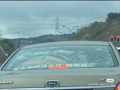 这是一张户外道路场景下的实拍静态图，画面主体是一辆吉利EC7汽车的尾部，汽车后窗张贴着包含脏话的标语“关上你的远光灯 哈麻批”，用于警示后方车辆关闭远光灯；背景可见阴天的天空、植被覆盖的山坡、高压输电塔、道路路灯以及交通信号灯，显示出这是在城市或城郊的道路环境中，画面右下角带有小红书平台的水印及对应用户的账号信息。