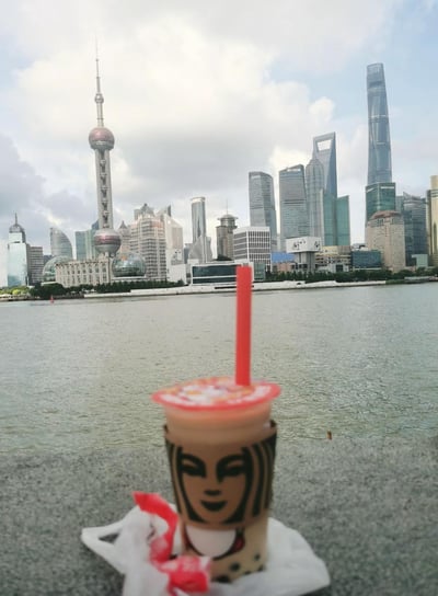 一张以上海浦东天际线为背景的照片，前景摆放着一杯奶茶。照片中可以清晰看到东方明珠电视塔、上海中心大厦等标志性建筑，展现了上海现代化的城市风貌。奶茶杯上有独特的人物面部图案设计，红色吸管直立，杯盖处可见奶茶配料。整体构图将日常饮品与城市地标结合，呈现出休闲惬意的氛围。