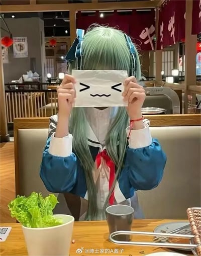 一位蓝绿色长发的coser坐在日式餐厅的座位上，双手举着一张画有简单表情（闭着的眼睛和波浪形嘴巴）的白色纸巾挡在脸前。她穿着蓝白相间的服装，领口系着红色蝴蝶结，背景中可以看到餐厅的木质桌椅、红色灯笼和带有日文汉字的红色布帘。桌子上摆放着一盆生菜和一个金属杯子，整体氛围轻松有趣。
