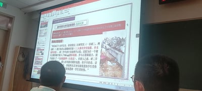 这是一张在室内教学场景中拍摄的投影屏幕翻拍图，屏幕展示的是实验室安全培训PPT里的「实验室事故案例分析」板块，聚焦2016年东华大学化学实验室爆炸事故。PPT详细介绍了事故发生的时间（2016年9月21日上午10点30分）、地点（上海松江大学园区东华大学化学化工与生物工程学院）、伤亡情况（两名学生重伤、一名学生轻伤），还追溯了事故过程：三名研究生未穿戴防护装备进行氧化石墨烯实验，在敞口容器中违规操作浓硫酸、石墨烯与高锰酸钾引发爆炸，同时配有事故后实验室一片狼藉的现场照片。画面下方可见两名听课人员的后脑勺，整体场景是在开展实验室安全警示教育。