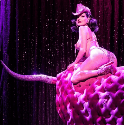 美国著名 burlesque 艺术家蒂塔·万提斯的舞台表演照。她身着闪亮粉色内衣套装与网袜，头戴同色系牛仔帽，侧身坐于巨大的粉色天鹅绒坐垫上，一条腿优雅伸展，鞋跟延伸出夸张的亮片长尾造型。背景是深紫色丝绒幕布，点缀着闪烁的星光效果，整体呈现出华丽复古的舞台氛围。