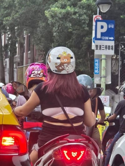 这是在台湾台北街头的车流中，一位紫发女生骑着摩托车的实拍场景。她的黑色短款上衣背后有特殊的镂空剪裁，看起来像是露出了浅紫色内衣，形成有趣的视觉错觉。她的头盔上贴着轻松熊（松弛熊）的卡通贴纸，周围还有其他骑乘摩托车的路人，背景里有指向「双连三停车场」的路牌，还有写着「阿彌陀佛」的标识，充满了台湾街头的日常通勤氛围。
这张图的趣味点在于上衣的设计造成的视觉错位，容易让人误以为女生的内衣外露，实际是衣服的特殊剪裁设计。