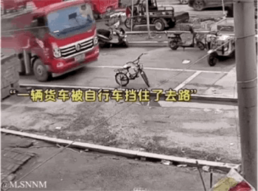 这是一张利用视觉错位效果的搞笑图片，画面展示了户外的一处场地，左侧停着一辆红色的大型东风货车，地面上的一辆自行车因为拍摄角度的问题，看起来正好位于货车的正前方，仿佛挡住了货车的去路。图中配有黄色的字幕文字：“一辆货车被自行车挡住了去路”，放大了这种错位带来的荒诞感，让小物体看似阻碍了大物体，制造出反差搞笑的效果。