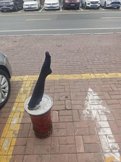 人行道的红砖地面上，一个红白相间的圆柱形柱子（疑似路障或小型灯柱）顶部，一只黑色袜子被直立放置，袜口朝上，形状宛如一条悬空的“小腿”。背景可见街道上停放的多辆汽车，左侧有部分银色轿车车身露出，整体场景充满生活中的意外趣味。