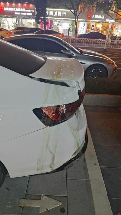 这是一张夜晚街头的实拍照片，画面主体是一辆停靠在人行道边缘的白色轿车，其车尾区域有明显的碰撞剐蹭损伤：尾灯、尾翼部分变形破损，并用透明胶带进行了临时固定，车身还残留着污渍痕迹。背景是一排临街商铺，能看到「金龙羽西化妆品商行」「青禾鞋行」「人间客店」等店铺招牌，周边还有其他停放的车辆，地面人行道上有一个指向左侧的箭头标识。
