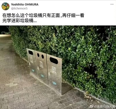 这张图展示了一个放置在绿植墙旁砖石路面上的金属垃圾桶，由于拍摄角度的巧妙巧合，垃圾桶的侧面与地面、绿植的位置关系让它看起来像是只有正面的平面板，形成了有趣的视觉错觉。配文调侃道“在想怎么这个垃圾桶只有正面，再仔细一看 光学迷彩垃圾桶....”，用“光学迷彩”来形容这个视角造成的奇特视觉效果，让普通的垃圾桶显得新奇又搞笑，充满了趣味性。