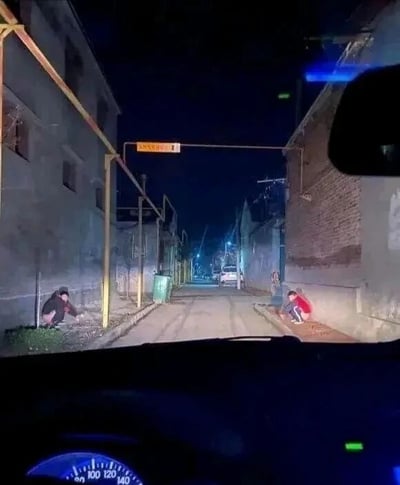 夜晚的狭窄小巷场景，从车内视角拍摄。巷子两侧是老旧的居民楼，左侧有一人蹲在墙边，右侧有两人（其中一人穿红色上衣）蹲坐于路边。巷子尽头停着一辆白色汽车，上方有黄色的街道指示牌，周围环境昏暗，仅有零星灯光照明，营造出静谧又略带神秘的氛围。