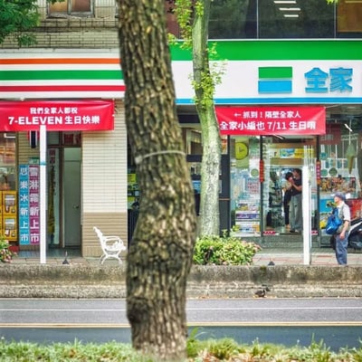 这张实拍街景图中，互为竞争对手的7-ELEVEN和全家两家连锁便利店相邻开设，分别在门店门口悬挂红色横幅进行趣味互动：左侧7-ELEVEN的横幅写有“我们全家人都祝7-ELEVEN生日快乐”，其中“全家人”一语双关，既指代本店内的所有员工，也玩了隔壁竞争对手“全家”便利店的品牌名称梗；右侧全家便利店打出横幅回应，内容为“←抓到！隔壁全家人 全家小编也7/11生日唷”，反过来玩7-ELEVEN的品牌名对应7月11日日期的梗，互动充满幽默感。画面前景有行道树，人行道上有一名背着蓝色背包的老年男性路人站立，全家门店门口还有两名顾客停留，整体是真实的街头场景，充满生活化的趣味。