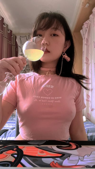 一位年轻女性在室内环境中手持酒杯饮酒的场景。她穿着粉色短袖T恤，上面印有白色文字和心形图案。脖子上戴着带有心形吊坠的choker项链，耳朵上佩戴着带有链条装饰的粉色耳环。她正将装有黄色液体的高脚杯举到嘴边饮用。背景中可以看到带有花纹的窗帘和室内陈设，整体氛围显得休闲舒适。