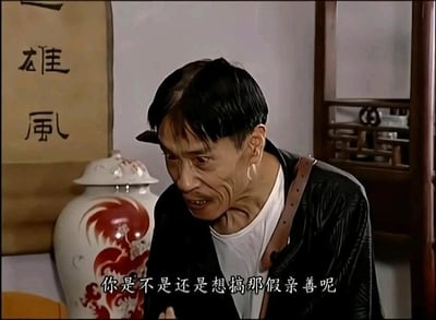 这是一张来自国产情景喜剧《地下交通站》的视频截图，画面主体是剧中角色贾贵，他头发凌乱稀疏，穿着黑色外褂搭配白色内搭，肩上挎着棕色背带。贾贵身体前倾，面部肌肉紧绷，眼神带着怀疑与质问的神色，表情刻薄又警惕，似乎正在近距离质问对方。画面下方配有白色字幕“你是不是还是想搞那假亲善呢”，背景是中式室内场景，左侧可见一幅写有“雄風”字样的书法作品，旁边摆放着一个带有红色龙纹图案的白色瓷罐，整体氛围充满了质问的张力，也贴合贾贵这个角色一贯的滑稽又刻薄的特质。