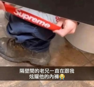 这张实拍图采用厕所隔间下方的拍摄视角，能看到相邻隔间的人正处于如厕状态，深蓝色裤子褪到了脚踝处，腰间露出带有潮牌Supreme红色白字标识的内裤腰边，图的下方配有中文配文“隔壁间的老兄一直在跟我炫耀他的内裤”，搭配流泪的表情符号，将他人正常如厕露出的内裤边戏谑解读为对方刻意炫耀，制造出强烈的反差搞笑效果。