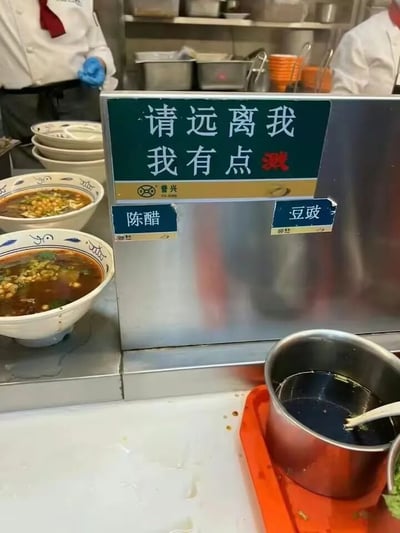 这是一张餐厅厨房的实拍照片，画面中心是一个不锈钢调料台，台面上立着一块绿色背景的搞笑提示牌，上面用白色大字写着“请远离我 我有点溅”，其中“溅”字用红色突出，巧妙利用谐音梗制造幽默效果（“溅”与“贱”谐音，既指调料可能溅出，又带调侃语气）。提示牌下方贴有蓝色标签，分别标注“陈醋”和“豆豉”。左侧摆放着几只青花瓷碗，里面盛有撒着葱花、黄豆等配料的深色汤品。背景中可见两位穿着白色厨师服、戴着蓝色手套的厨师正在忙碌，后方货架上整齐叠放着不锈钢盆和橙色碗碟，整体呈现出真实忙碌的后厨氛围。
