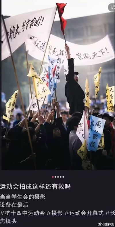 这张照片拍摄的是杭十四中运动会开幕式场景，学生们高举写有各类激昂标语的旗帜，其中一名学生站在人群中高高举起红旗，周围环绕着写满书法文字的黄色、白色旗帜，场面看起来十分热血沸腾。照片下方配有文字“运动会拍成这样还有救吗 当当学生会的摄影 设备在最后”，并带有相关话题标签。整体画面通过长焦镜头压缩感，营造出了极具视觉冲击力的效果，将普通的校园运动会拍出了史诗级的氛围感。