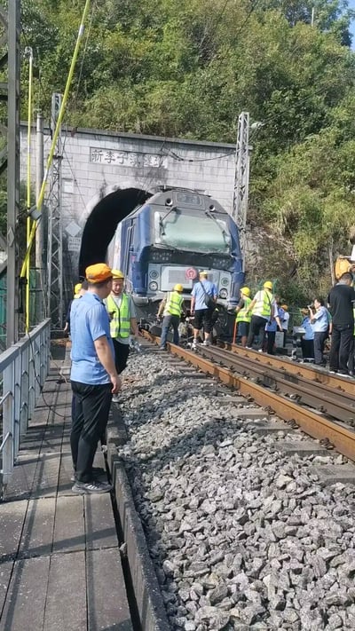 图片中一列蓝色火车正从标有“新李子溪隧道”的隧道口缓缓驶出，隧道上方是茂密的绿色山林。铁轨两侧及轨道上有多名身着蓝色工作服、黄色安全背心和黄色安全帽的施工人员，部分人靠近火车头，似乎在进行检查或维护工作。轨道旁堆放着碎石，上方有金属支架和电线，整体场景展现了铁路工程现场的日常作业状态。
