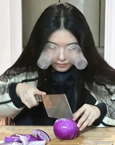 图中展示了一位长发女性正在切紫色洋葱的场景，为了避免切洋葱时释放的刺激性气体刺激眼部导致流泪，她想出了一个简易的防护办法：将两个透明塑料杯分别罩在双眼部位，以此阻挡刺激性气体接触眼睛。她手持菜刀，正专注地处理案板上的洋葱，案板上还摆放着几片已经切好的洋葱块，整个画面呈现出一种生活趣味小妙招的实践场景，既实用又充满创意。
