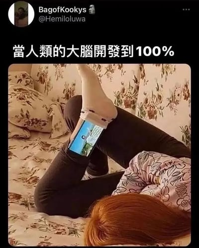 这张梗图展示了一个人以不寻常的姿势躺在床上，双腿抬起，用脚夹着手机观看。图片上方配有文字“當人類的大腦開發到100%”（当人类的大脑开发到100%），通过幽默的方式调侃了即使人类大脑完全开发，人们可能仍然会用这种慵懒的方式使用手机。