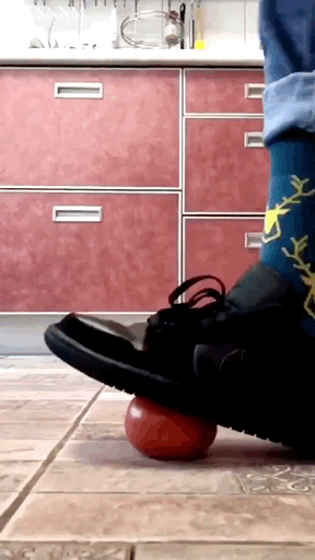 这是一段厨房场景下的动态GIF，画面中穿着黑色皮鞋、带有黄色树枝图案蓝色袜子的脚踩在一颗红色小球上，试图用脚将球碾碎，但小球在脚下不断滚动，看起来是徒劳的尝试，充满了滑稽搞笑的氛围。图中没有双关、谐音梗或暗示内容。