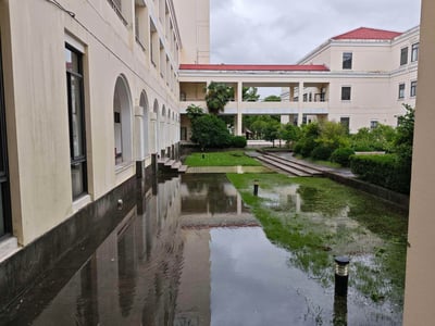 这张图片展示了雨后校园或机构建筑的庭院景象。画面中央是一个被积水覆盖的庭院，水面平静如镜，清晰倒映出周围的建筑轮廓。庭院两侧是米白色的古典风格建筑，左侧建筑带有连续的拱形门廊，右侧建筑则为平顶设计。远处可以看到连接两栋建筑的红色屋顶走廊，以及庭院中修剪整齐的绿色草坪和各种灌木、树木。天空阴沉，云层较厚，显示出刚下过雨的天气特征。整个场景呈现出一种宁静而略带压抑的氛围，积水的庭院为原本整洁的校园增添了几分特别的景致。