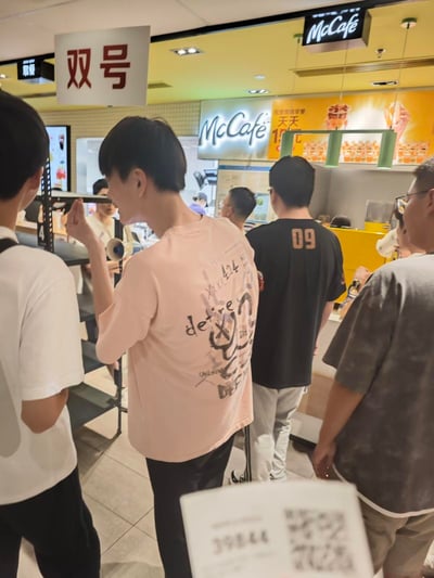 一张在麦当劳餐厅内拍摄的照片，展示了顾客在McCafé柜台前排队的场景。画面中可以看到几位顾客的背影，其中一位穿着粉色T恤的年轻人尤为显眼，T恤背面印有"desire"字样及图案。柜台上方悬挂着醒目的"McCafé"标志和促销海报，左侧有一个标有"双号"的指示牌，可能用于引导顾客排队或分流。整体环境明亮，呈现出典型的快餐店氛围。
