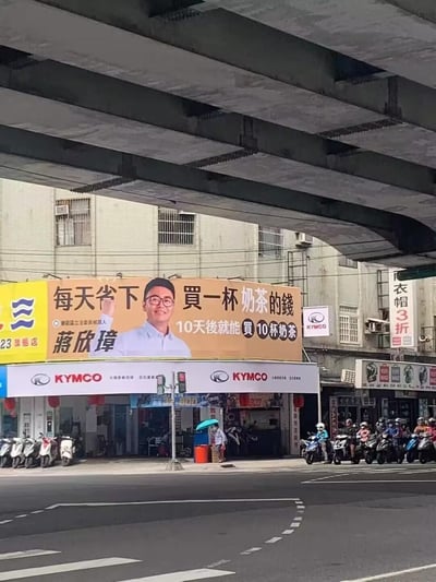 城市街道场景下，一座大型广告牌格外醒目，上面有戴着眼镜的男子蒋欣璋微笑挥手。广告牌主打标语“每天省下买一杯奶茶的钱，10天后就能买10杯奶茶”，逻辑看似矛盾却引人发笑。下方是KYMCO摩托车店，街道上停放着多辆摩托车，有行人打着伞经过，展现出真实的城市街景。