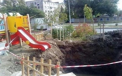 图片展示了一个户外游乐场场景，核心元素为一个红黄相间的儿童滑梯（黄色平台搭配红色滑道），滑梯右侧紧邻一个巨大的不规则土坑，坑深约1-2米，周围用红白相间的警示带简单围拢。背景可见多层居民楼、绿色树木及街道，整体环境显示为居民区附属的游乐设施区域。土坑边缘泥土松散，与滑梯距离极近，存在明显的安全隐患，可能是未完成的施工工程或意外塌陷形成。