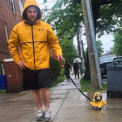 A man wearing a bright yellow rain jacket with the hood up and black shorts is walking a small dog on a leash. The dog is also dressed in a matching yellow rain jacket, creating an adorable coordinated look. They're on a wet sidewalk in a residential neighborhood with brick buildings, green trees, and other pedestrians in the background, suggesting a rainy day. The man has a friendly smile, adding to the heartwarming vibe of the scene.