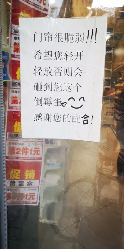 这是一张拍摄于线下饮品售卖店铺（从促销贴纸判断为售卖怡宝水的便利店或小型超市）门帘处的照片。门帘上用透明胶带粘贴着一张白色纸张，上面打印着趣味提示语：“门帘很脆弱！！！希望您轻开轻放否则会砸到您这个倒霉蛋😊感谢您的配合！”，用幽默自嘲的语气提醒顾客小心对待门帘，避免门帘损坏或砸到顾客。图片左侧下方有红色的促销贴纸，标注“第2件1元”“促销 怡宝水”，活动时间为2023年7月4日至7月17日，门帘为透明塑料材质，整体场景是店铺入口处。