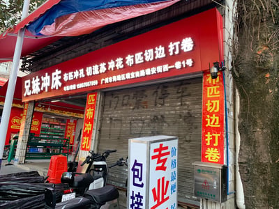 这是一张实拍的广州街边店铺照片，展示了位于广州市海珠区瑞宝南路瑞安街西一巷1号的「兄妹冲床」布料加工店。店铺红色招牌醒目，标注了主营项目：布匹冲孔、切流苏、冲花、布匹切边打卷，同时留有联系电话。店铺卷帘门处于关闭状态，门口停放着小牛电动的电动车，旁边立着写有「专业 价格最便 包接」的宣传牌，还有一个电动汽车充电箱，背景中可见售卖槟榔的店铺，整体呈现出充满生活气息的市井街边场景。
无双关、谐音梗或暗示类内容。