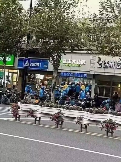 这是一张城市街头的实拍照片，画面中心是益丰大药房的门店，店门前聚集了大量身着蓝色统一制服的外卖骑手，他们或站在原地，或低头查看手机，身旁停放着多辆外卖电动车。药房两侧是其他沿街商铺，左侧是带有“JINSON”标识的店铺，右侧是“舒适精选”店铺，街道边缘设置了带有绿植的花箱，上方有行道树遮挡部分视线。整个场景展现了城市外卖配送从业者的工作日常，没有双关、谐音梗或其他暗示内容。